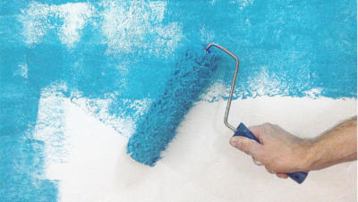 A close-up of a hand and blue paint roller applying paint to a white wall.
